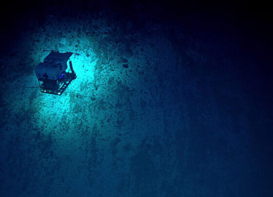 A photo of a remotely operated vehicle exploring the Mariana Trench. It appears as a cube hovering over a lighter blue patch surrounded by darkness.