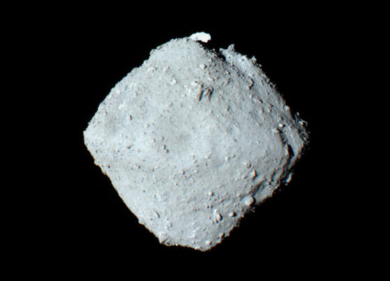 A photo take of the asteroid Ryugu. A light gray circular shape in the center on a deep black background.