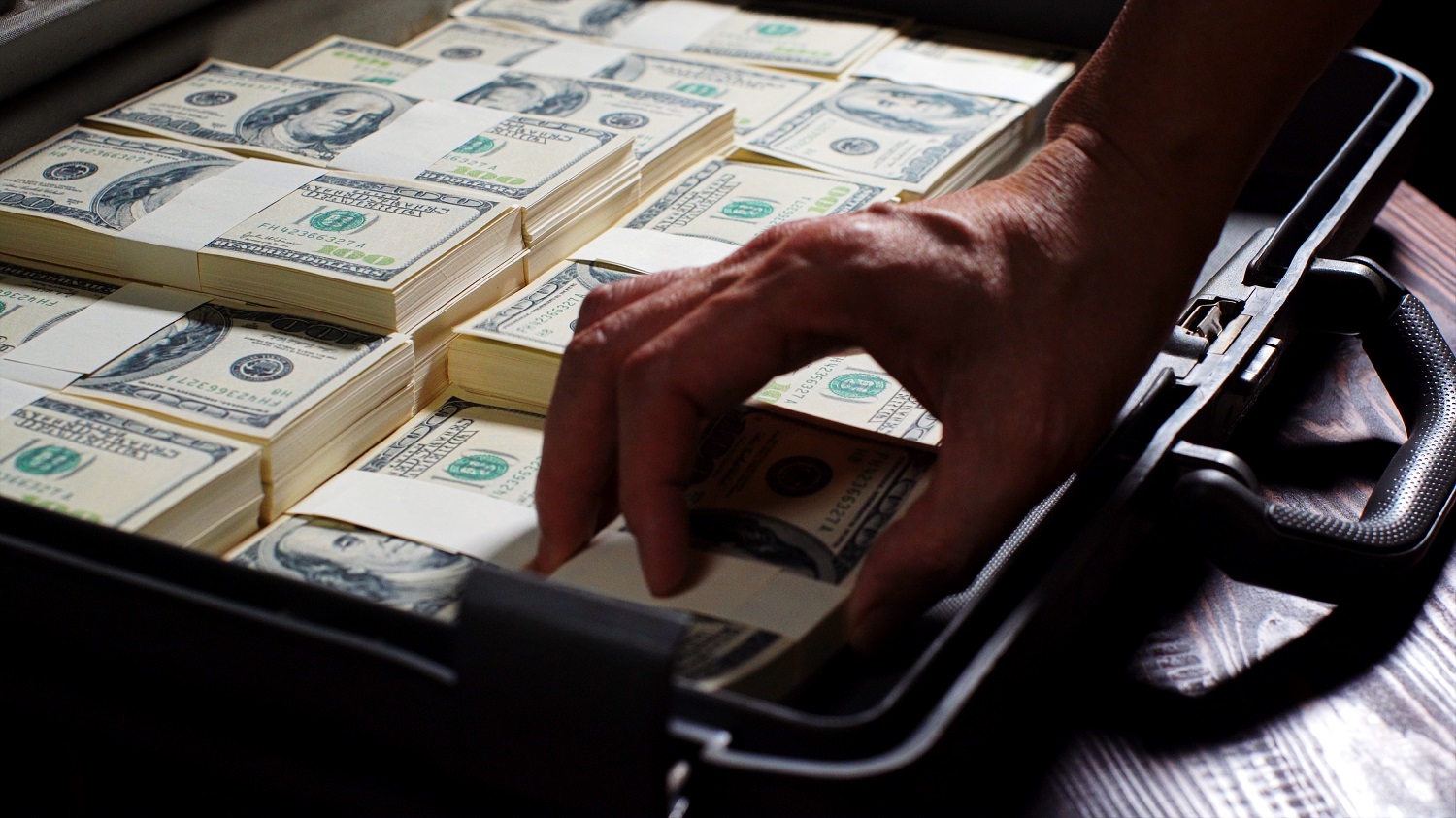 A person&rsquo;s hand grabs a pile of US dollar bills from a suitcase filled with bundles of cash.