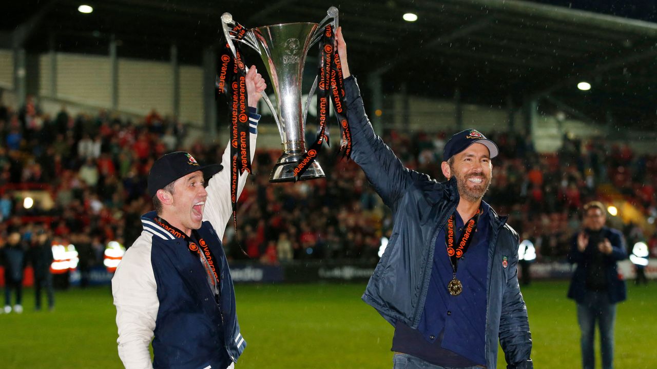 Rob McElhenney and Ryan Reynolds celebrate with the trophy on the pitch.