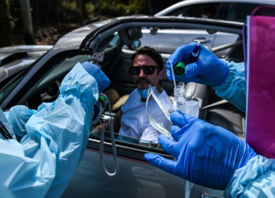 People wearing gloves and full-body personal protective equipment prepare a COVID-19 test for a person sitting in a car wearing sunglasses