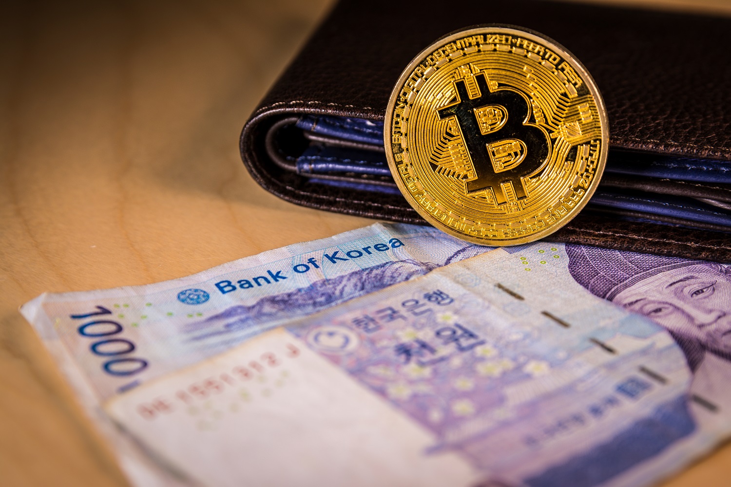 A leather wallet rests on a table, on top of South Korean banknotes. A metal token intended to represent a Bitcoin token leans against the wallet.