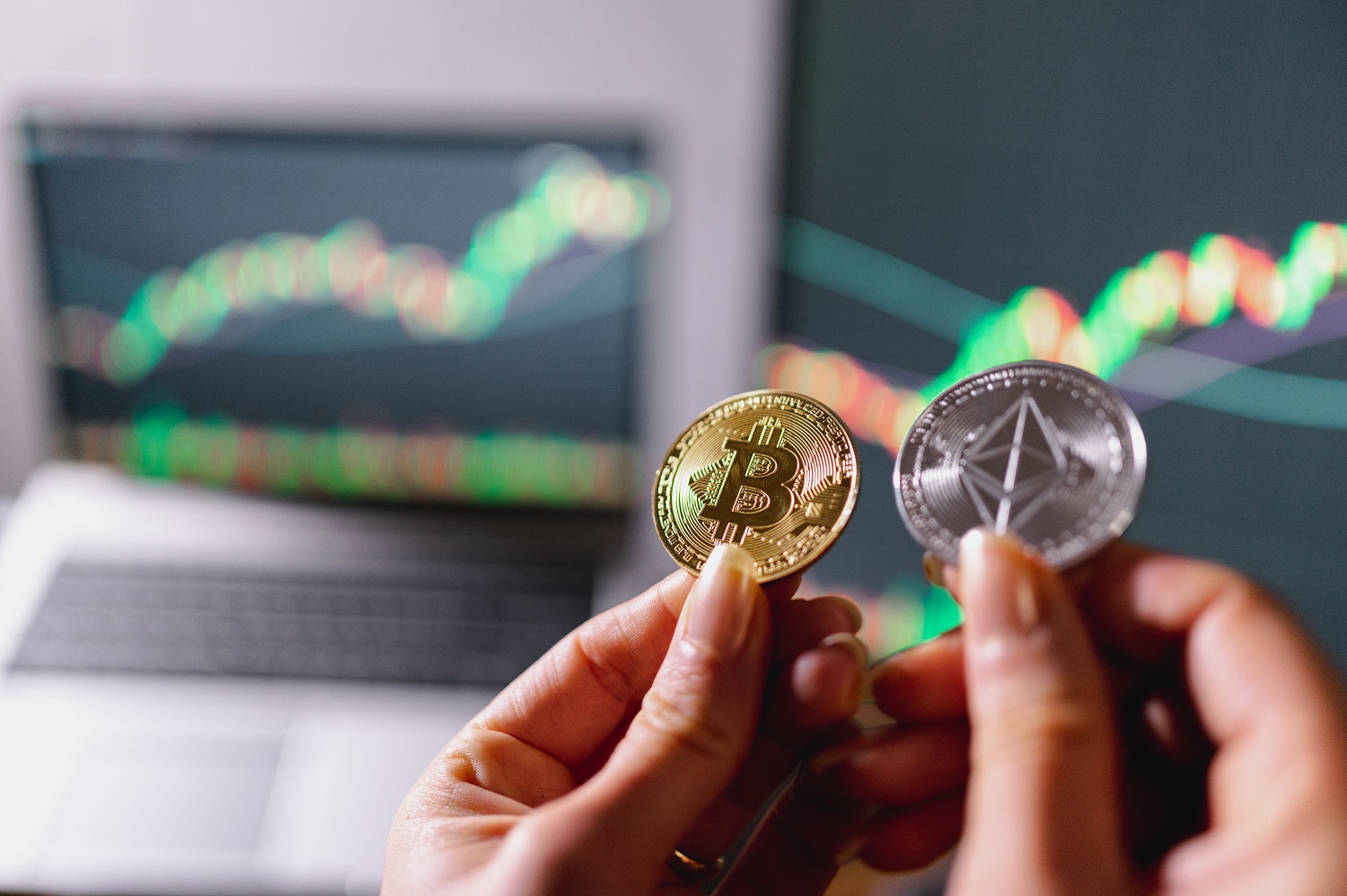 A woman’s hands hold metal tokens intended to represent Ethereum and Bitcoin coin with a laptop displaying a graph in the background.