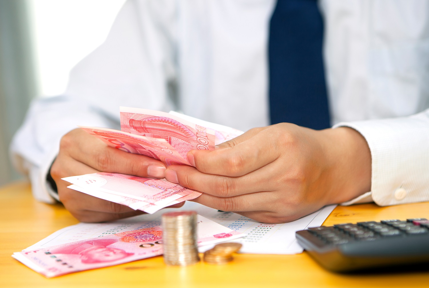 A man in a shirt and tie, seated at a table, counts Chinese banknotes, with a pile of coins also resting on the table.