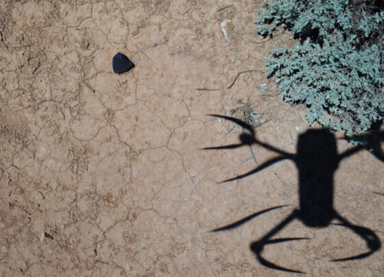 A drone casts a shadow on the ground while it hovers above a dark object flagged as a potential meteorite.