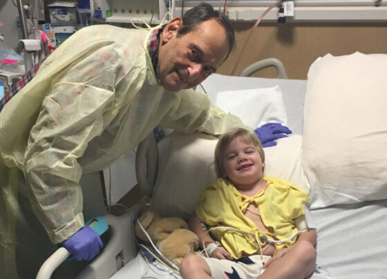 Will Ungerer lies in a hospital bed, next to a teddy bear, as doctor Jerry Mendell leans over him. Both are looking at the camera and smiling.