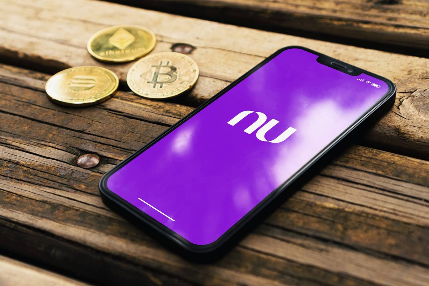 The Nubank bank logo on a smartphone screen on a table next to metal tokens intended to represent popular cryptoassets.