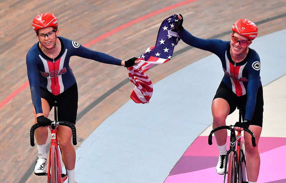 Two women smiles while riding bicycles, holding an American flag between them.