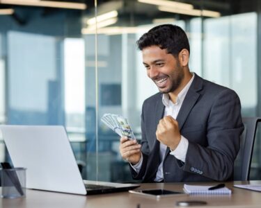 A smiling investor, holding money in one hand, looks at something on a laptop worth getting excited about.