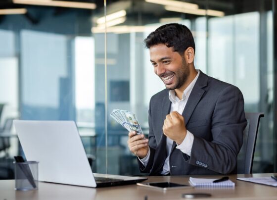 A smiling investor, holding money in one hand, looks at something on a laptop worth getting excited about.